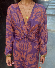 Abito elegante viola con motivo arancione, scollo a V profondo, maniche lunghe, indossato da donna. Accessori: collana dorata, bracciali.