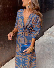 Donna con abito lungo blu a motivi dorati, clutch blu, orecchini pendenti e bracciali dorati. Moda elegante, abbigliamento femminile di tendenza.