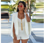 Donna con occhiali da sole specchiati, indossa un elegante completo bianco con mantella e collana lunga dorata, sfondo urbano con palme.