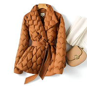 Giacca trapuntata marrone con cintura, appesa su gruccia, accanto a borsa beige e sciarpa bianca. Moda autunno inverno, abbigliamento donna elegante.