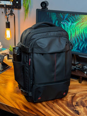 Zaino nero da viaggio su scrivania in legno, con tasche laterali e dettagli rossi, ideale per laptop e accessori tecnologici. Ambiente ufficio moderno.