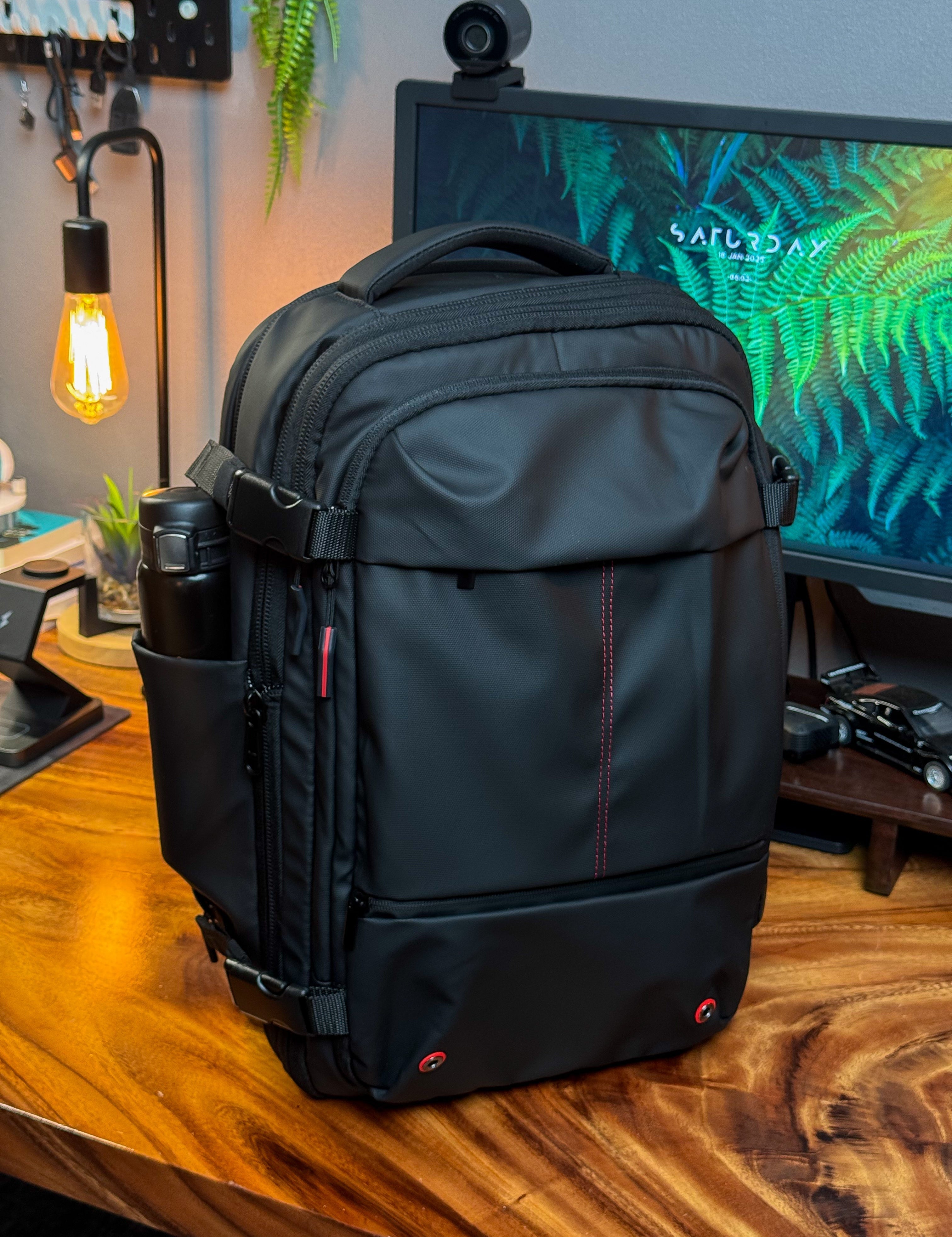 Zaino nero da viaggio su scrivania in legno, con tasche laterali e dettagli rossi, ideale per laptop e accessori tecnologici. Ambiente ufficio moderno.