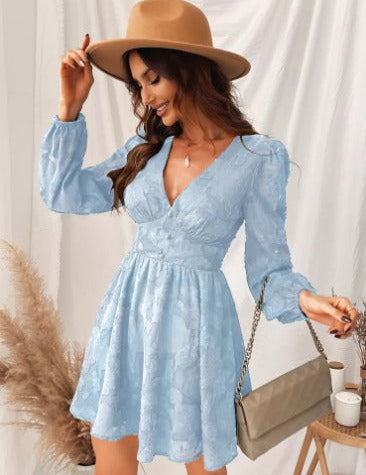 Abito corto in pizzo azzurro con maniche lunghe, indossato da donna con cappello beige. Accessori: borsa a tracolla beige. Elegante e alla moda.