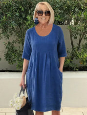 Donna con abito blu in lino, occhiali da sole, orecchini blu, borsa nera con fiori bianchi. Sfondo di piante verdi. Moda estiva casual.