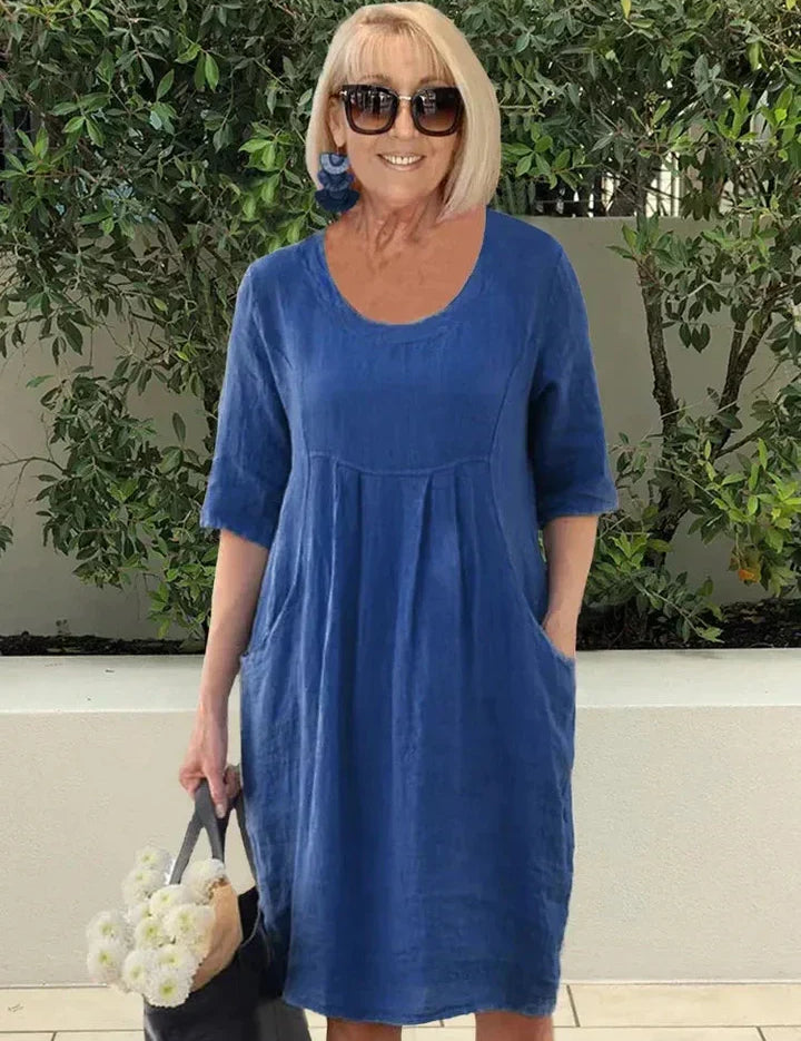 Donna con abito blu in lino, occhiali da sole, orecchini blu, borsa nera con fiori bianchi. Sfondo di piante verdi. Moda estiva casual.