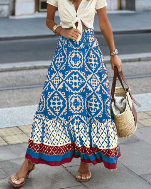 Donna con gonna lunga boho a motivi geometrici blu e bianchi, camicetta bianca annodata, sandali marroni e borsa di paglia. Moda estiva casual.