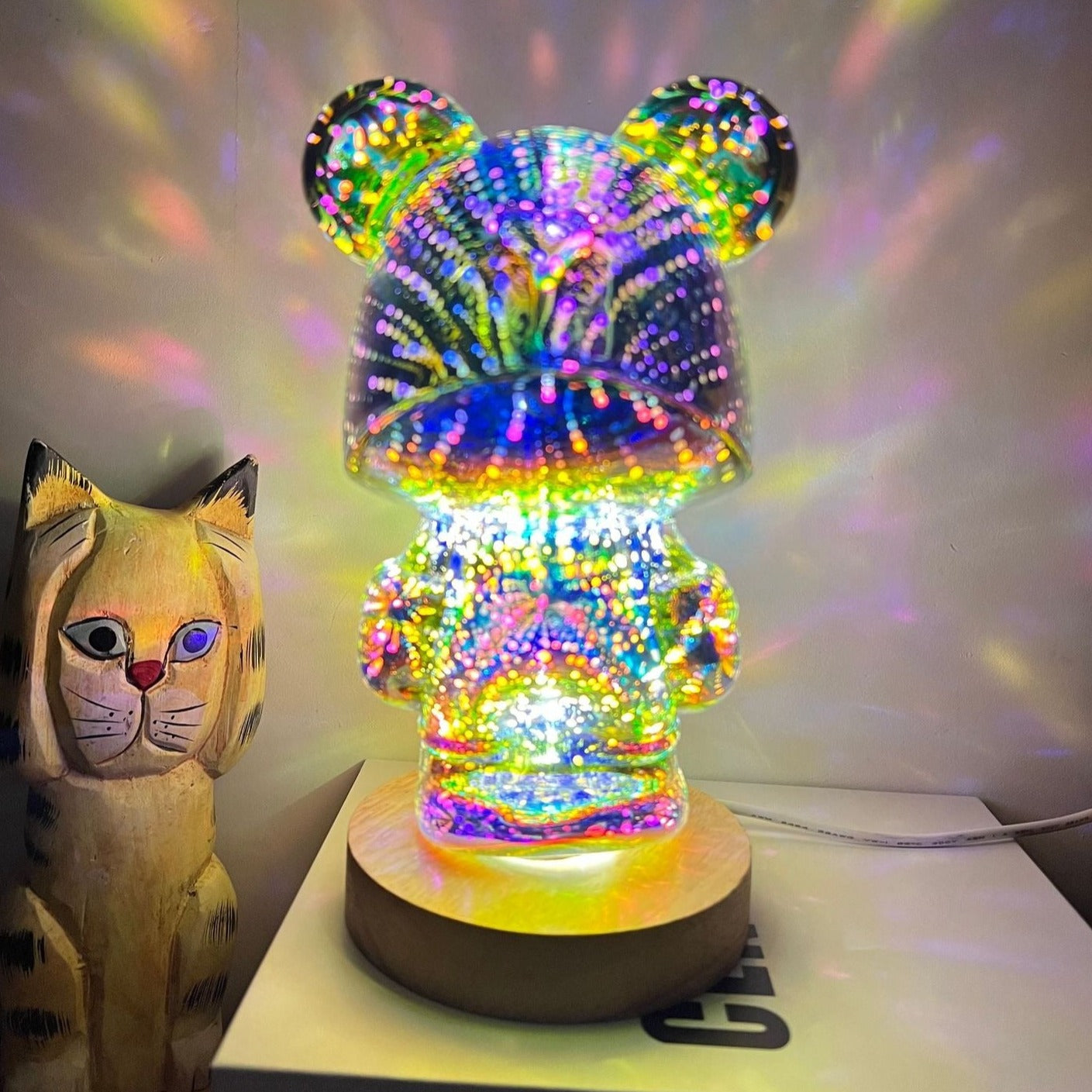 Lampada LED a forma di orso con effetto 3D multicolore su base in legno, accanto a una statuetta di gatto in legno. Decorazione moderna e luminosa.