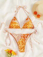 Bikini arancione con stampa boho, top a triangolo e slip con laccetti, su sfondo bianco. Accessori: cappello di paglia, occhiali da sole, fiori gialli.