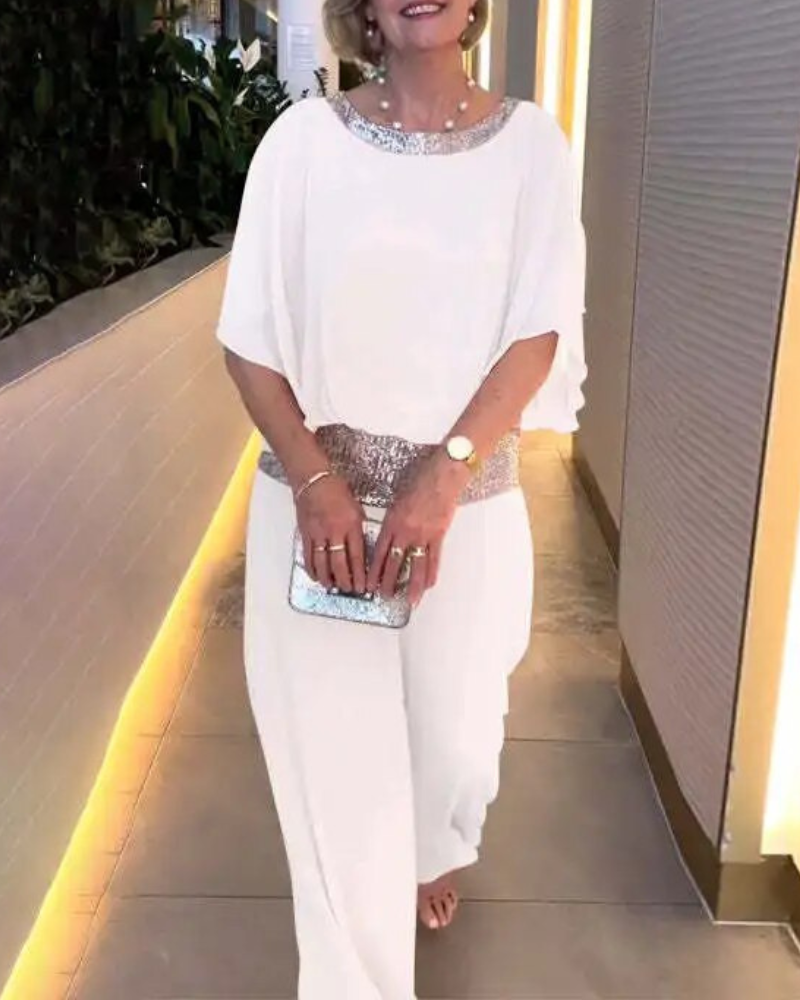 Donna in elegante tuta bianca con dettagli scintillanti, clutch argentata, orecchini pendenti, e orologio dorato. Look da sera sofisticato.