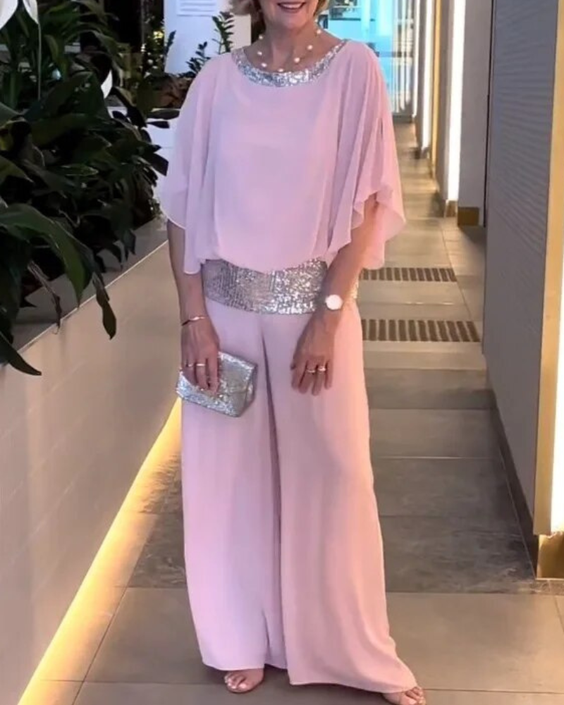 Donna in elegante tuta rosa con dettagli scintillanti, pochette argento, in corridoio moderno. Abbigliamento da sera chic e sofisticato.