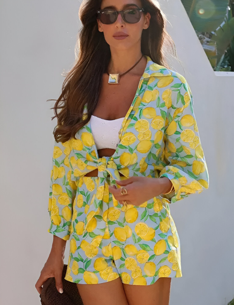 Donna con occhiali da sole indossa un completo estivo con stampa di limoni, giacca e pantaloncini, su sfondo bianco. Moda estiva, abbigliamento casual.