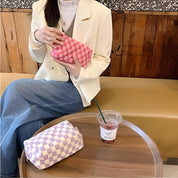 Donna con giacca bianca tiene pochette a scacchi rosa, seduta accanto a borsa a scacchi viola su tavolo in legno. Moda casual, accessori eleganti.