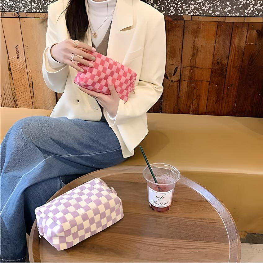 Donna con giacca bianca tiene pochette a scacchi rosa, seduta accanto a borsa a scacchi viola su tavolo in legno. Moda casual, accessori eleganti.