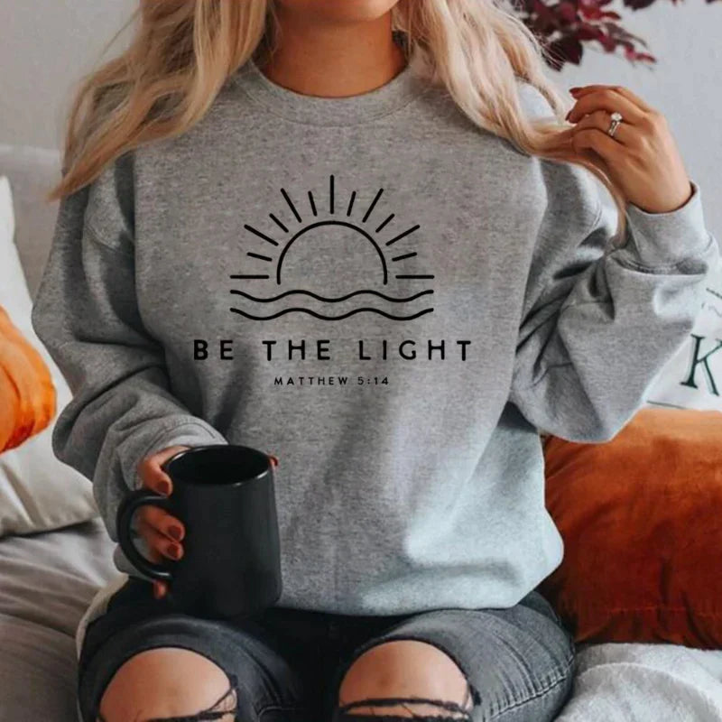 Donna seduta con felpa grigia "Be the Light", sole stilizzato, versetto biblico Matteo 5:14, tazza nera in mano. Moda casual, abbigliamento ispirato.