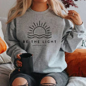 Donna seduta con felpa grigia "Be the Light", sole stilizzato, versetto biblico Matteo 5:14, tazza nera in mano. Moda casual, abbigliamento ispirato.