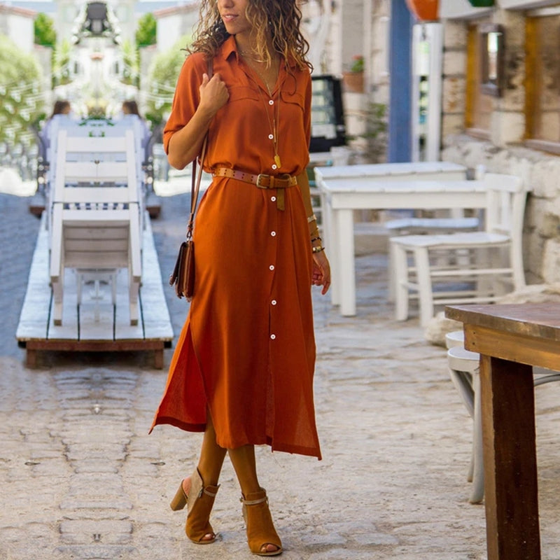 Donna con abito lungo arancione, cintura marrone e sandali, cammina in una strada di ciottoli all'aperto. Moda estiva, abbigliamento casual elegante.