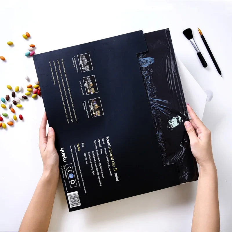 Mani che tengono un poster da grattare nero con skyline, accanto a caramelle colorate e pennelli. Kit fai-da-te artistico, regalo creativo.