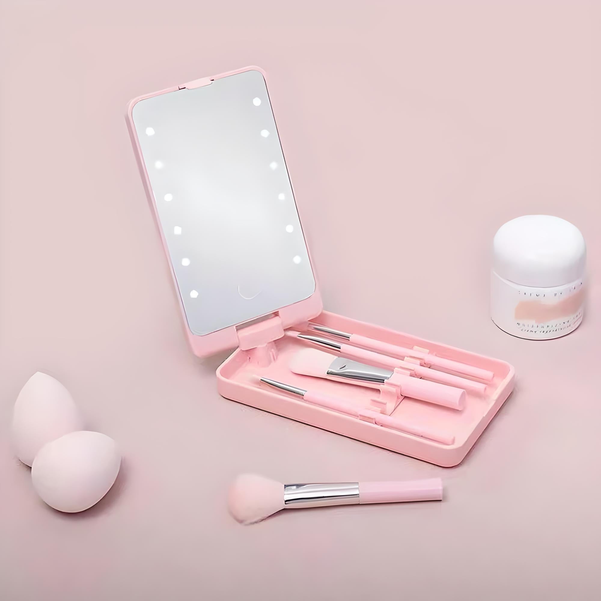 Set trucco rosa con specchio illuminato, pennelli, spugne e crema viso su sfondo rosa. Kit cosmetico portatile per make-up professionale.