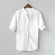 Camicia bianca in lino da uomo, maniche corte, colletto alla coreana, appesa su gruccia di legno, sfondo grigio. Elegante e casual.