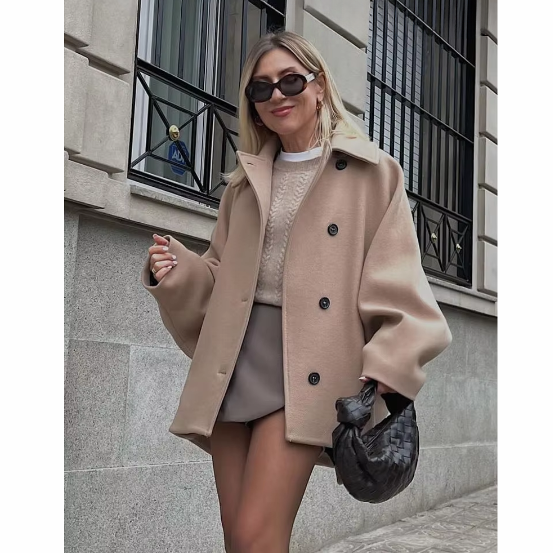 Donna con cappotto beige oversize, occhiali da sole neri e borsa intrecciata nera, posa davanti a edificio urbano. Moda autunnale elegante.