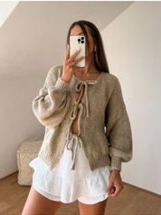 Donna con cardigan beige a maglia con lacci e maniche a sbuffo, abbinato a shorts bianchi a balze. Outfit moda autunnale casual chic.