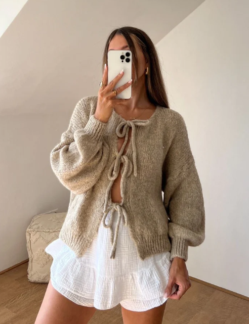 Donna con cardigan beige a maglia con lacci e maniche a sbuffo, abbinato a shorts bianchi a balze. Outfit moda autunnale casual chic.