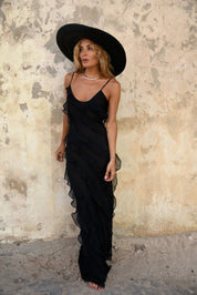 Donna con abito lungo nero elegante e cappello a tesa larga, posa contro un muro rustico. Moda estiva, abbigliamento femminile chic.