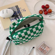 Astuccio a scacchi verde e bianco su letto, circondato da libri, pennelli trucco e piattino con ciliegie. Accessorio moda, organizzazione cosmetici.