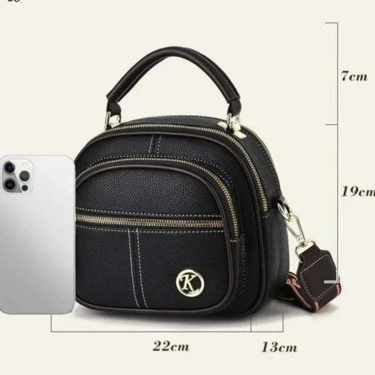 Borsa a tracolla nera in pelle PU con logo "K", tasca frontale con zip, dimensioni 22x13x19 cm, accanto a uno smartphone per confronto.