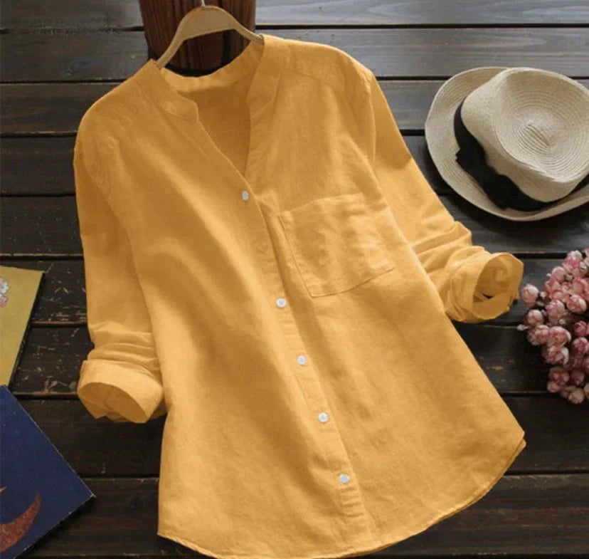 Camicia donna gialla in lino, maniche lunghe, colletto alla coreana, bottoni bianchi, su tavolo di legno con cappello e fiori rosa.