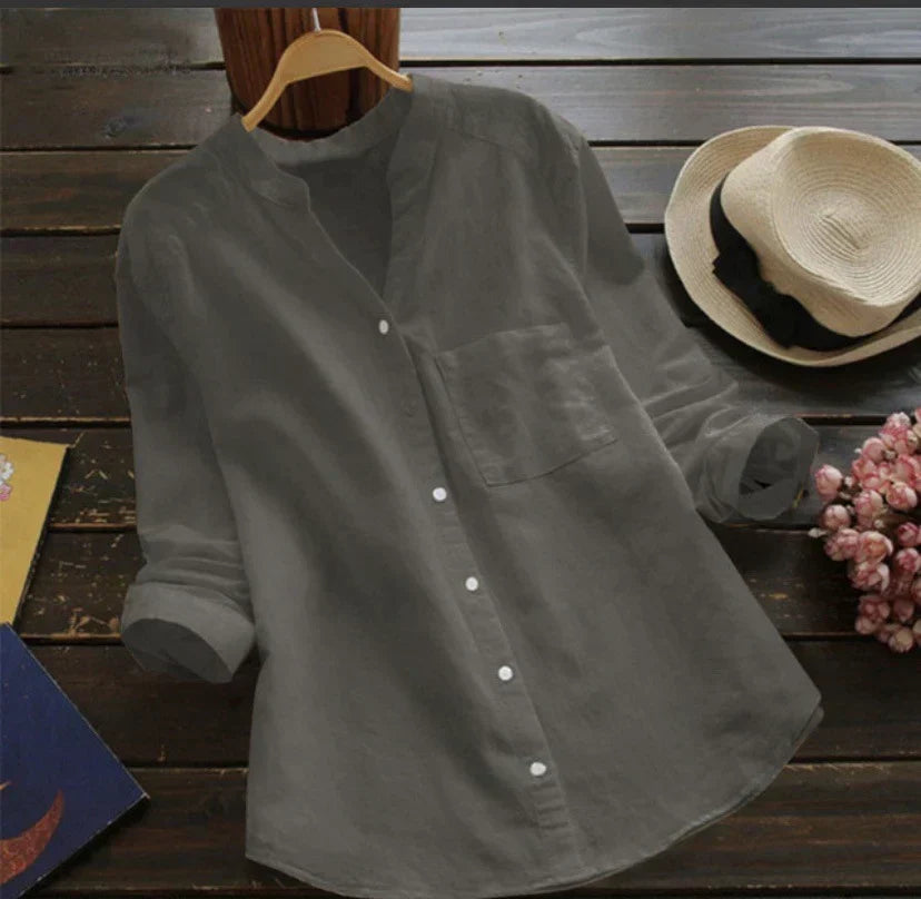 Camicia grigia in lino su appendiabiti, stile casual, con colletto alla coreana e maniche lunghe, accanto a cappello di paglia e fiori rosa.