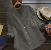 Camicia grigia in lino su appendiabiti, stile casual, con colletto alla coreana e maniche lunghe, accanto a cappello di paglia e fiori rosa.