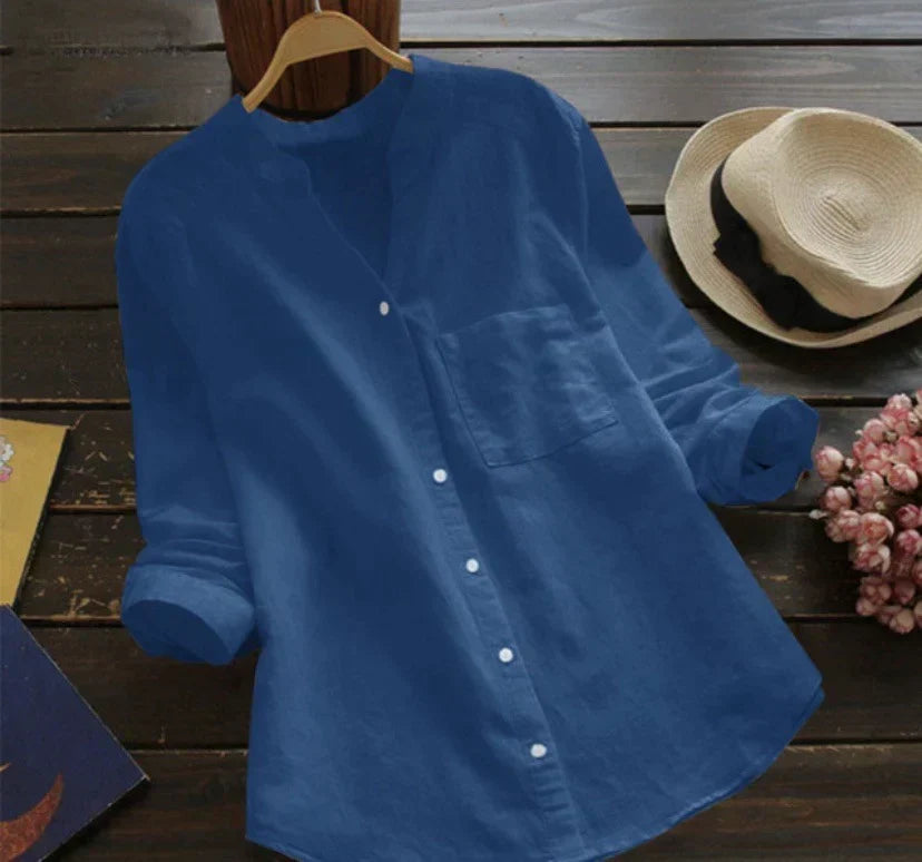 Camicia blu da donna in cotone, maniche lunghe, colletto alla coreana, bottoni frontali, su sfondo di legno con cappello di paglia e fiori rosa.