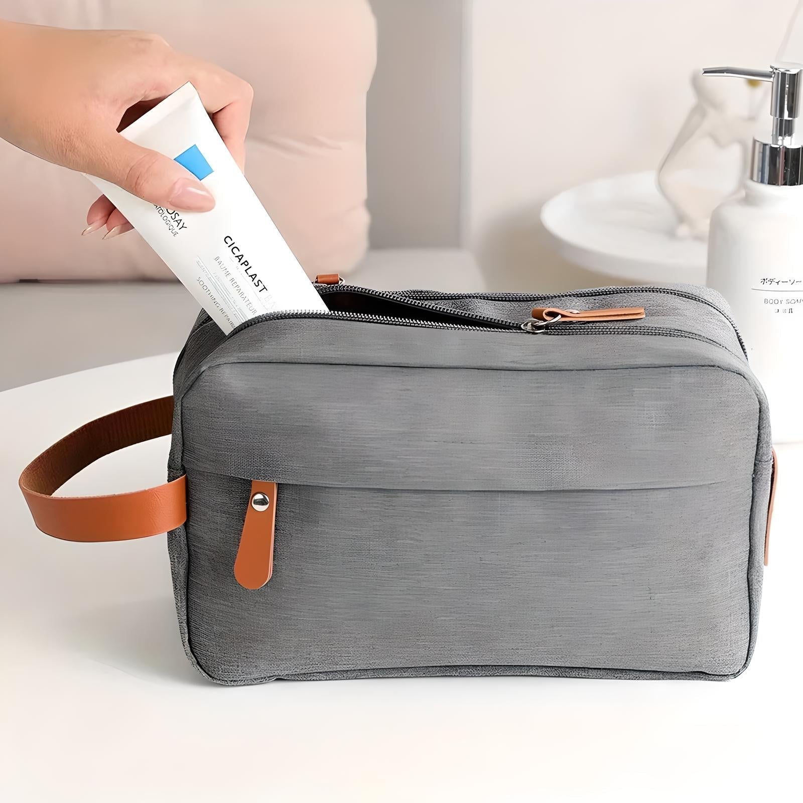 Borsa da toilette grigia con manico in pelle marrone, tasca frontale e zip, contenente un tubetto di crema. Accessorio da viaggio elegante e funzionale.