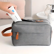 Borsa da toilette grigia con manico in pelle marrone, tasca frontale e zip, contenente un tubetto di crema. Accessorio da viaggio elegante e funzionale.
