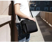 Uomo con borsa a tracolla nera, design moderno, indossata su spalla destra. Ideale per lavoro e viaggio. Sfondo urbano.
