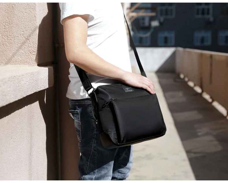 Uomo con borsa a tracolla nera, design moderno, indossata su spalla destra. Ideale per lavoro e viaggio. Sfondo urbano.