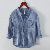 Camicia di lino blu da uomo, maniche lunghe, colletto alla coreana, tasca frontale, design casual, appesa su gruccia di legno.