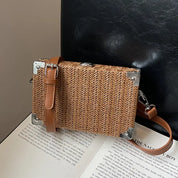 Borsa a tracolla in paglia intrecciata con angoli rinforzati in metallo e cinturino in pelle marrone, posata su un libro aperto. Elegante e versatile.