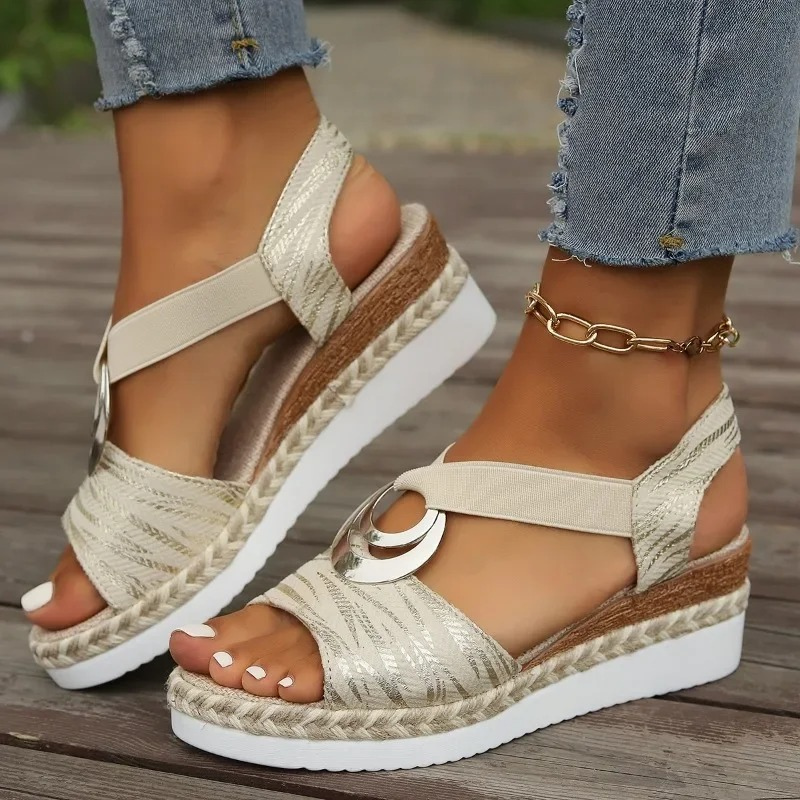 Sandali estivi da donna con zeppa, cinturino beige, dettagli metallici, suola in gomma bianca, abbinati a jeans strappati e cavigliera dorata.