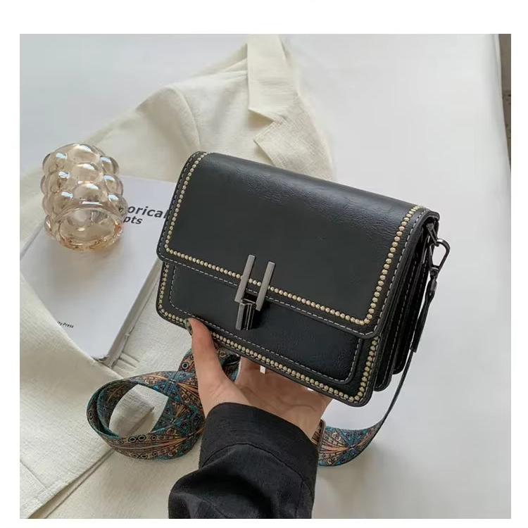 Borsa a tracolla nera in pelle con dettagli borchiati, chiusura a fibbia, su giacca beige. Accessorio moda elegante e versatile.