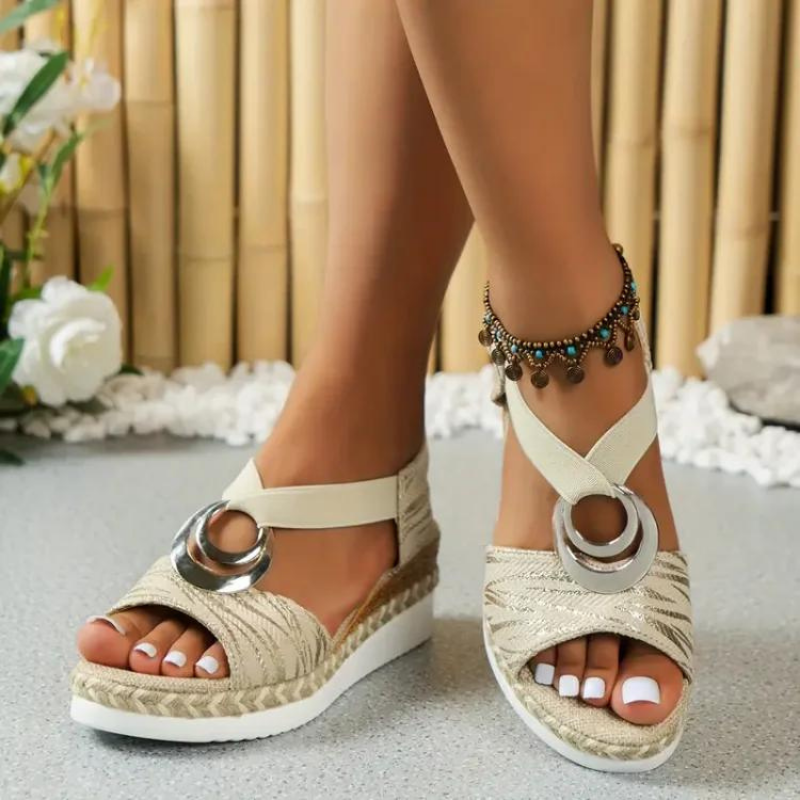Sandali estivi da donna con zeppa, colore beige, decorati con anello metallico e cinturino alla caviglia, su sfondo di bambù e fiori bianchi.