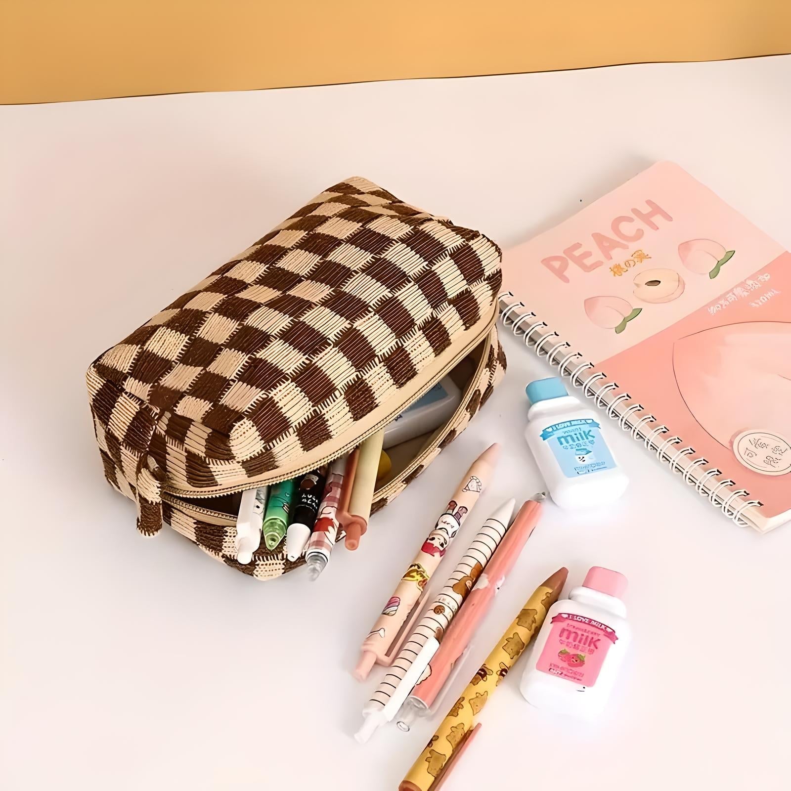 Astuccio a scacchi marrone con penne colorate, quaderno rosa "Peach" e bottigliette di latte su scrivania bianca. Accessori scolastici eleganti.