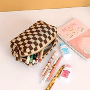 Astuccio a scacchi marrone con penne colorate, quaderno rosa "Peach" e bottigliette di latte su scrivania bianca. Accessori scolastici eleganti.
