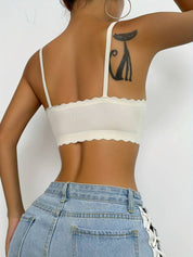Top corto bianco con spalline sottili e bordo ondulato, indossato da una donna con jeans a vita alta. Tatuaggio di gatto nero visibile sul braccio.