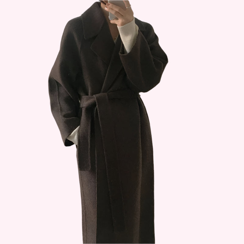 Cappotto lungo marrone scuro in lana, con cintura in vita, indossato su maglione bianco. Elegante abbigliamento invernale per donna.
