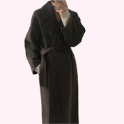 Cappotto lungo marrone scuro in lana, con cintura in vita, indossato su maglione bianco. Elegante abbigliamento invernale per donna.