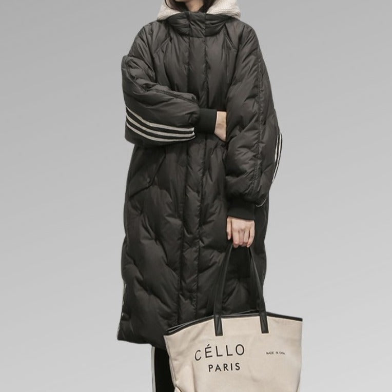 Cappotto lungo imbottito nero con cappuccio, dettagli a righe sulle maniche, abbinato a borsa beige "Céllo Paris". Moda invernale elegante.