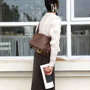 Donna con maglione bianco e gonna marrone, borsa a tracolla marrone, fermaglio per capelli, davanti a finestra. Stile autunnale elegante.