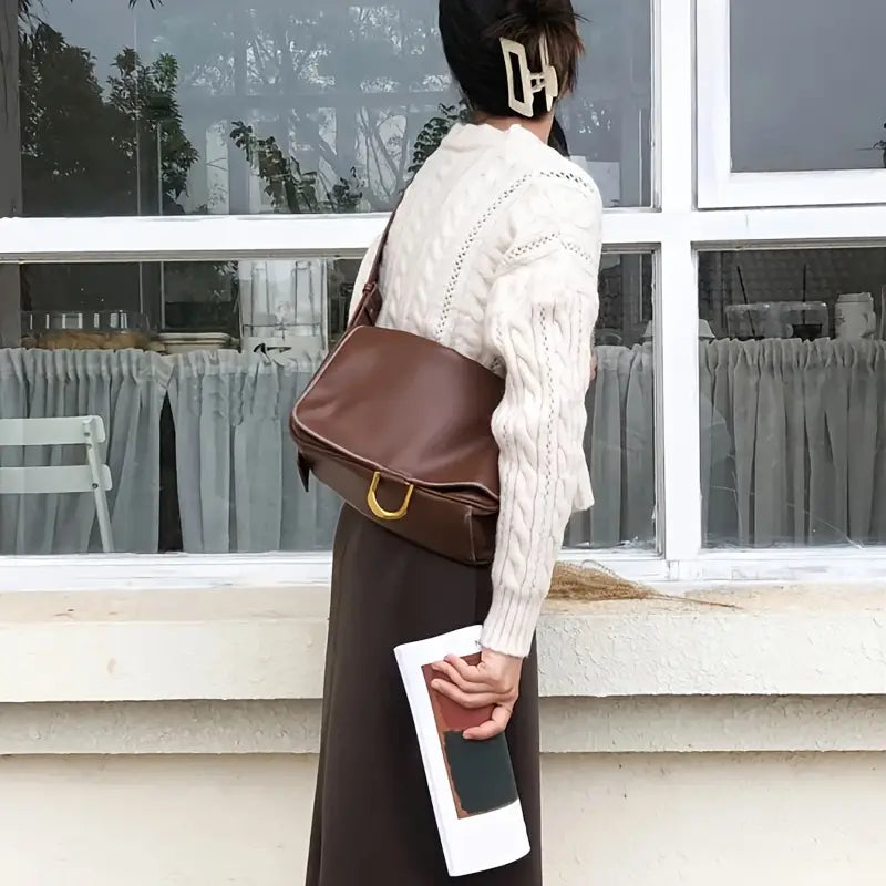 Donna con maglione bianco e gonna marrone, borsa a tracolla marrone, fermaglio per capelli, davanti a finestra. Stile autunnale elegante.